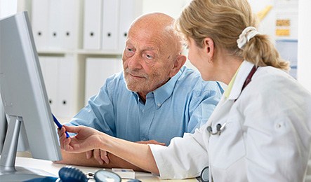 En kvinnlig läkare pekar på en datorskärm och en äldre man sitter bredvid och tittar på datorskärmen. Foto: iStock 